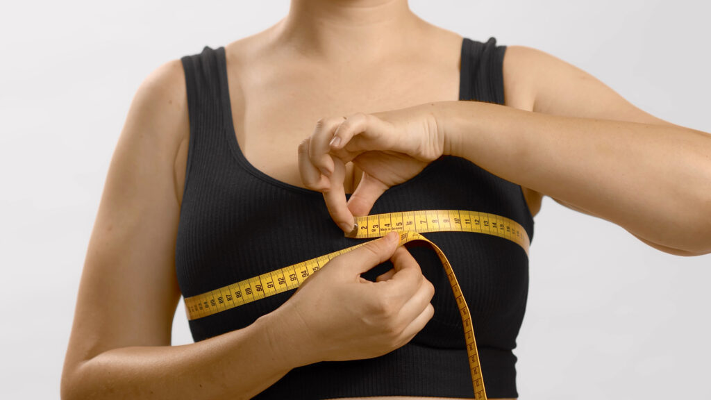 Woman wearing a black tank top measures her bust with a yellow measuring tape to determine her correct bra size. Dr Tracy Pfeifer - NEW YORK